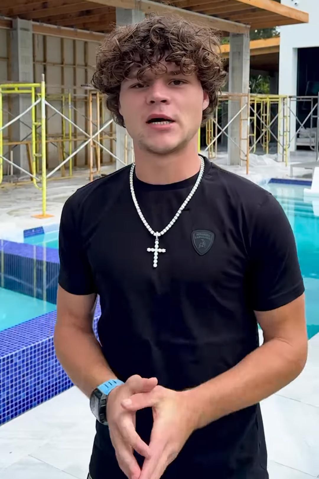 Jack Doherty, a young man with curly brown hair, wearing a black t-shirt and a diamond cross necklace, standing by a pool with a construction site in the background.