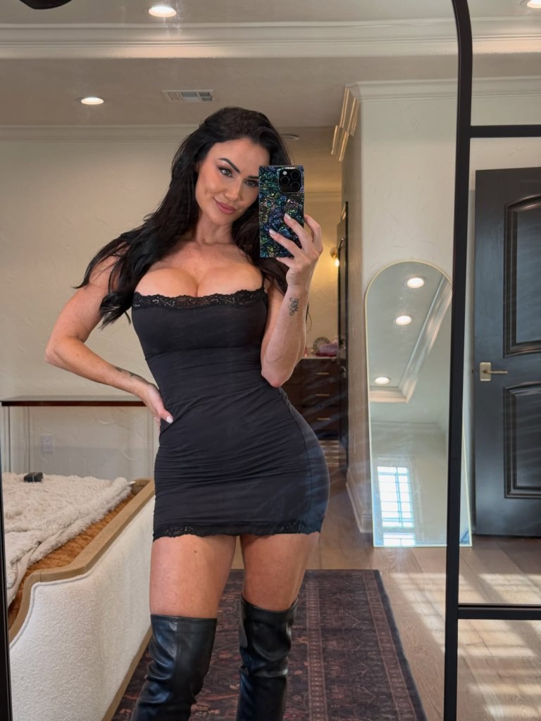 A woman in a black mini-dress and boots taking a mirror selfie.