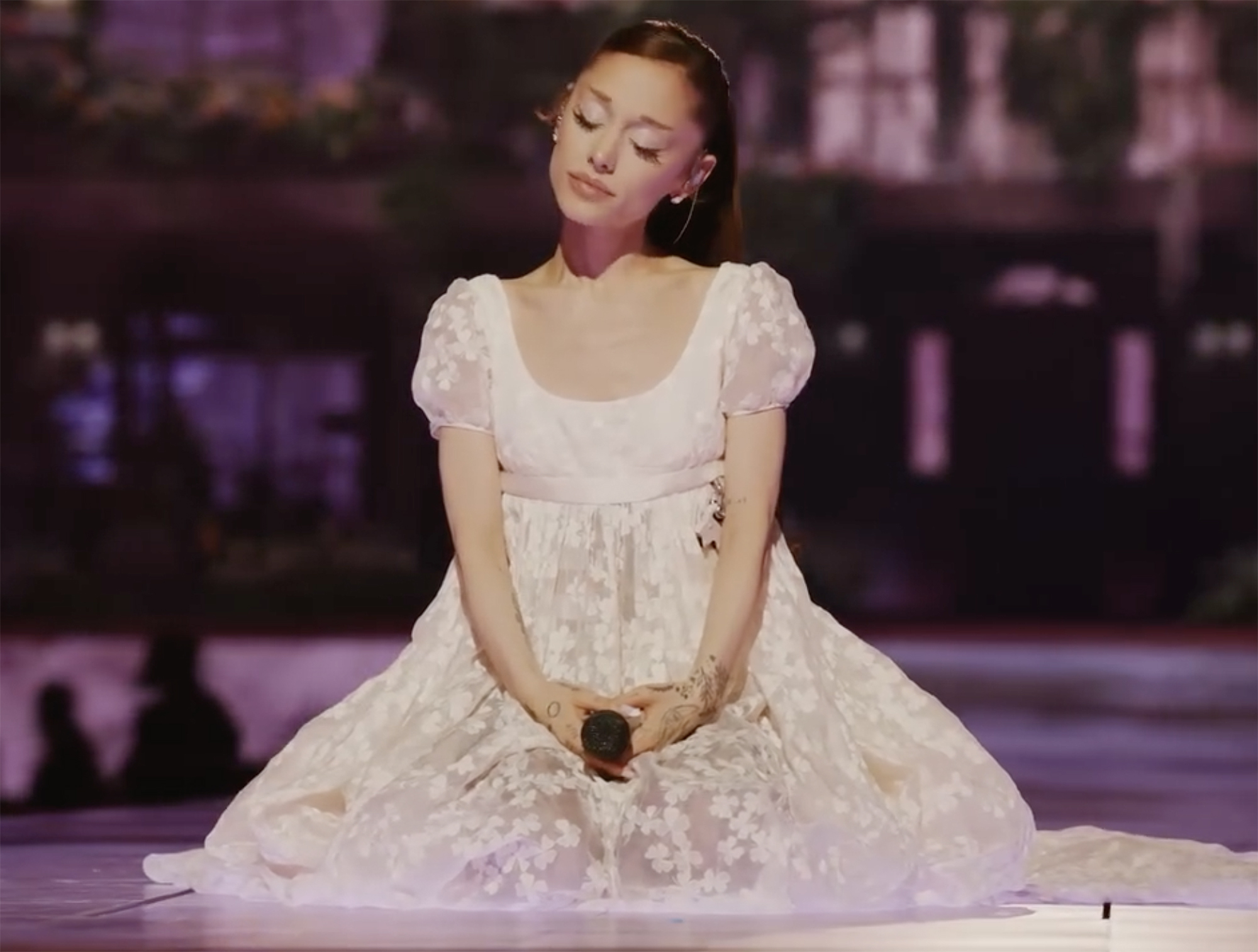 Ariana Grande wearing a white floral dress and holding a microphone, seated on stage.