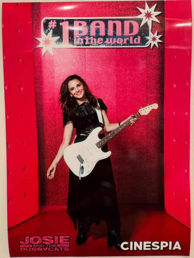 Rachael Leigh Cook holding a white electric guitar in front of a sparkly pink background with 