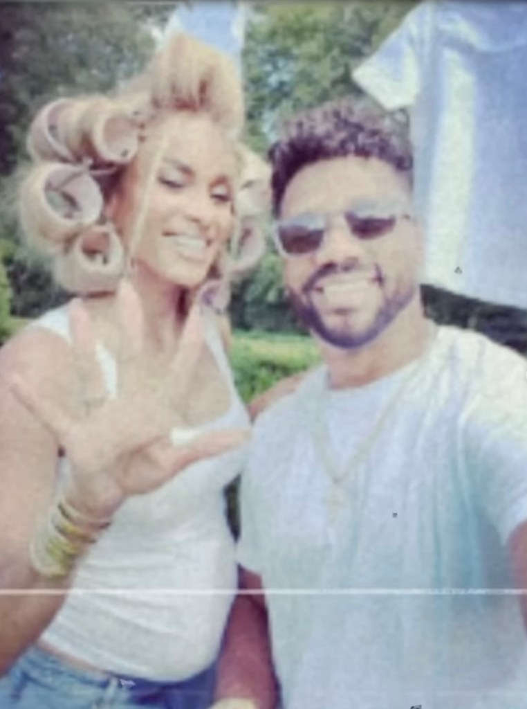 Ciara and Russell Wilson in a selfie, Ciara revealing her pregnancy while holding up four fingers, with a baby onesie hanging in the background.