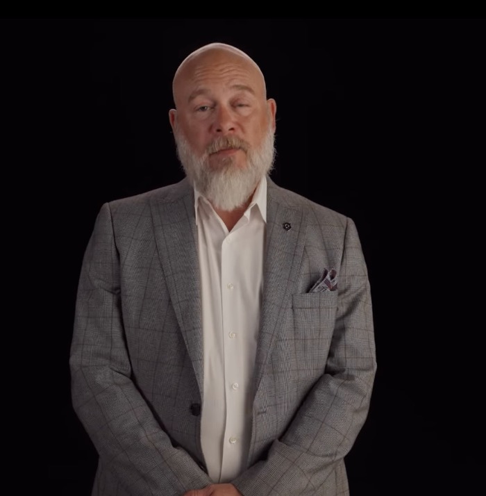 A bald man with a white beard wearing a suit and white shirt, against a black background.