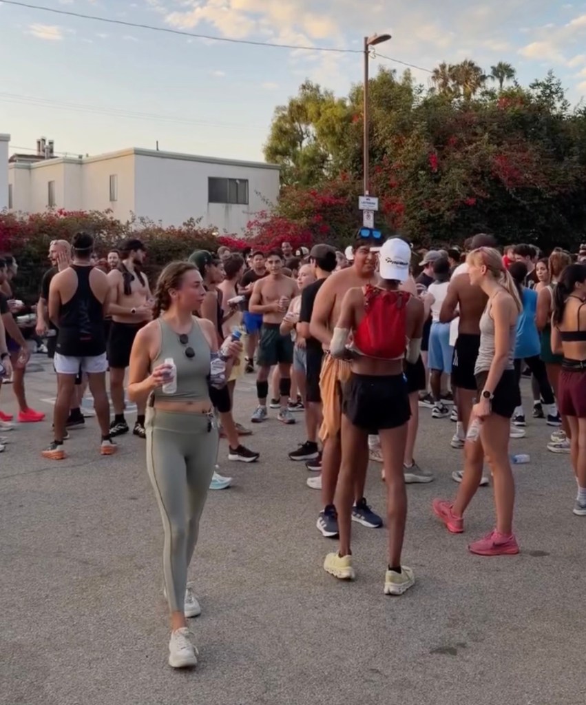 A crowd of people, many shirtless, in athletic wear, gathered outdoors in what appears to be a street or parking lot for the Venice Beach run club in California.