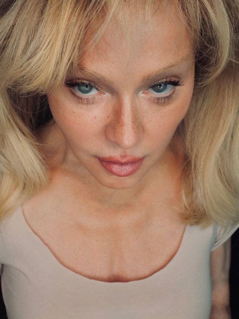Madonna looking up at the camera, wearing a light-colored top, with blonde hair, visible freckles, and striking blue eyes.