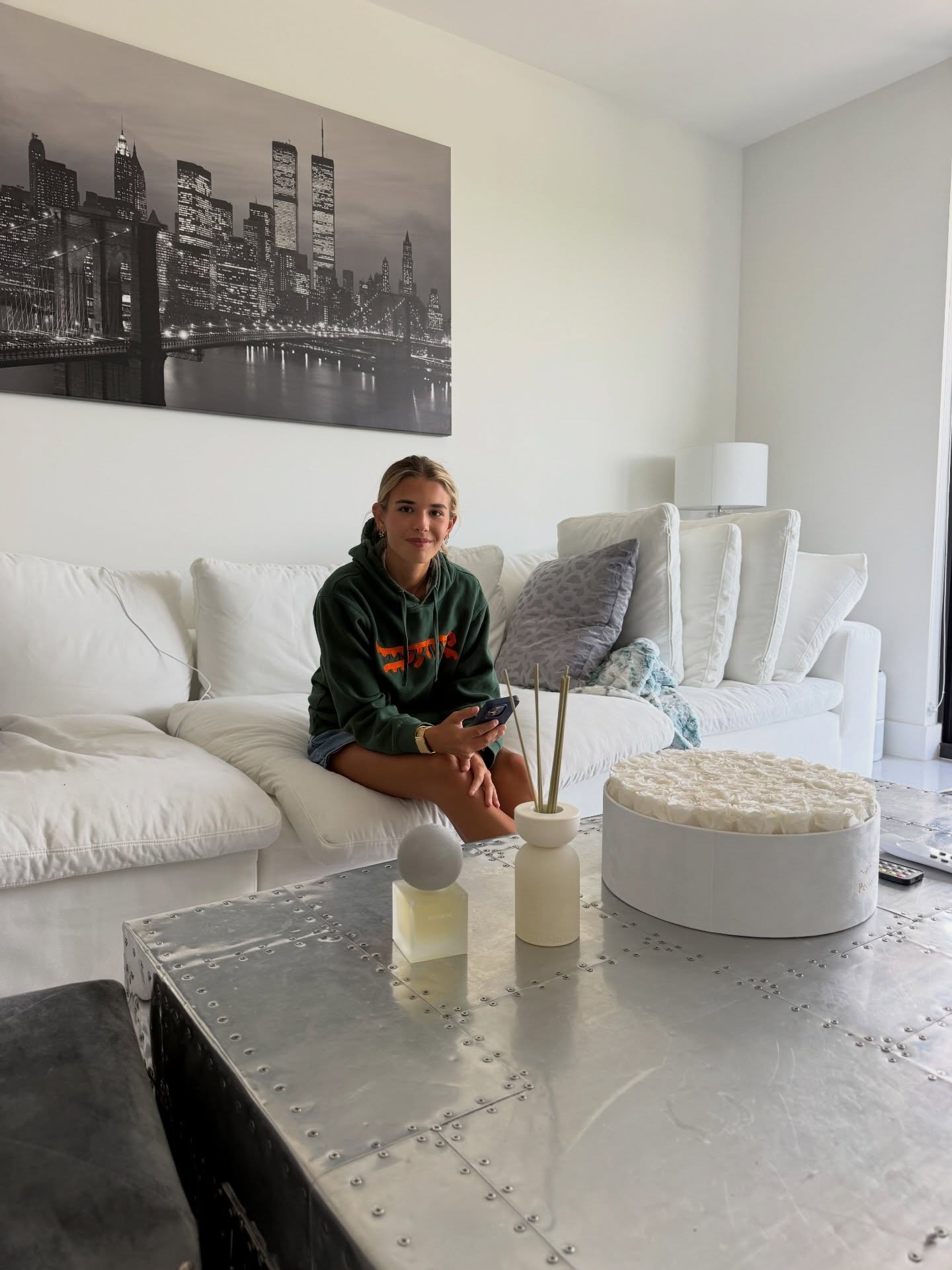A young woman in a green hoodie sits on a white couch, holding a phone, with a large black and white city artwork behind her.