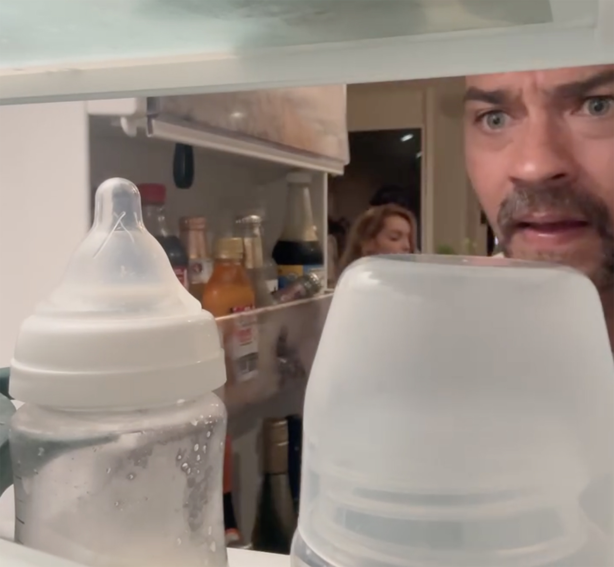 Jesse Williams peeking over baby bottles in a refrigerator, appearing startled.
