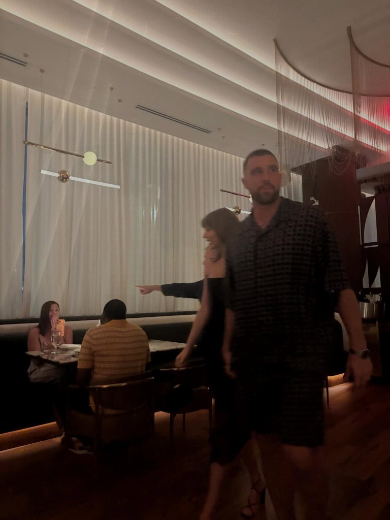 Taylor Swift and Travis Kelce walking and holding hands in a dimly lit restaurant.