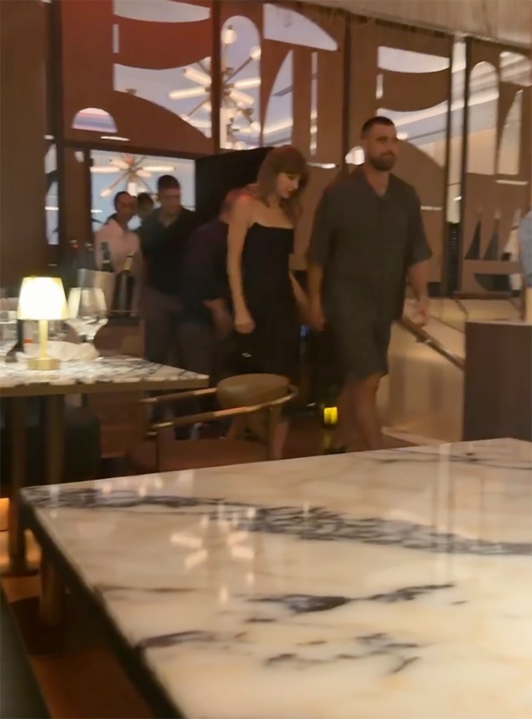 Taylor Swift and Travis Kelce hold hands while walking through a restaurant.