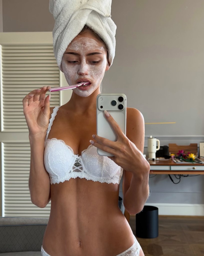 Woman in a white lace bra, face mask, and hair wrapped in a towel brushing her teeth while taking a mirror selfie.