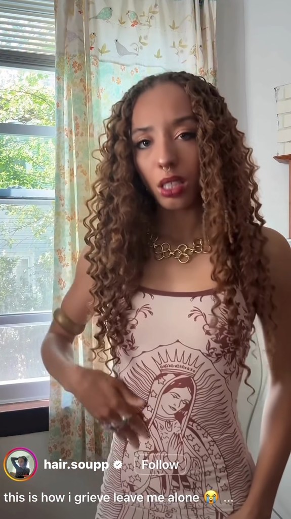 A person with long, curly hair wearing a pink dress with an illustration of the Virgin Mary, a gold necklace, and a nose ring.