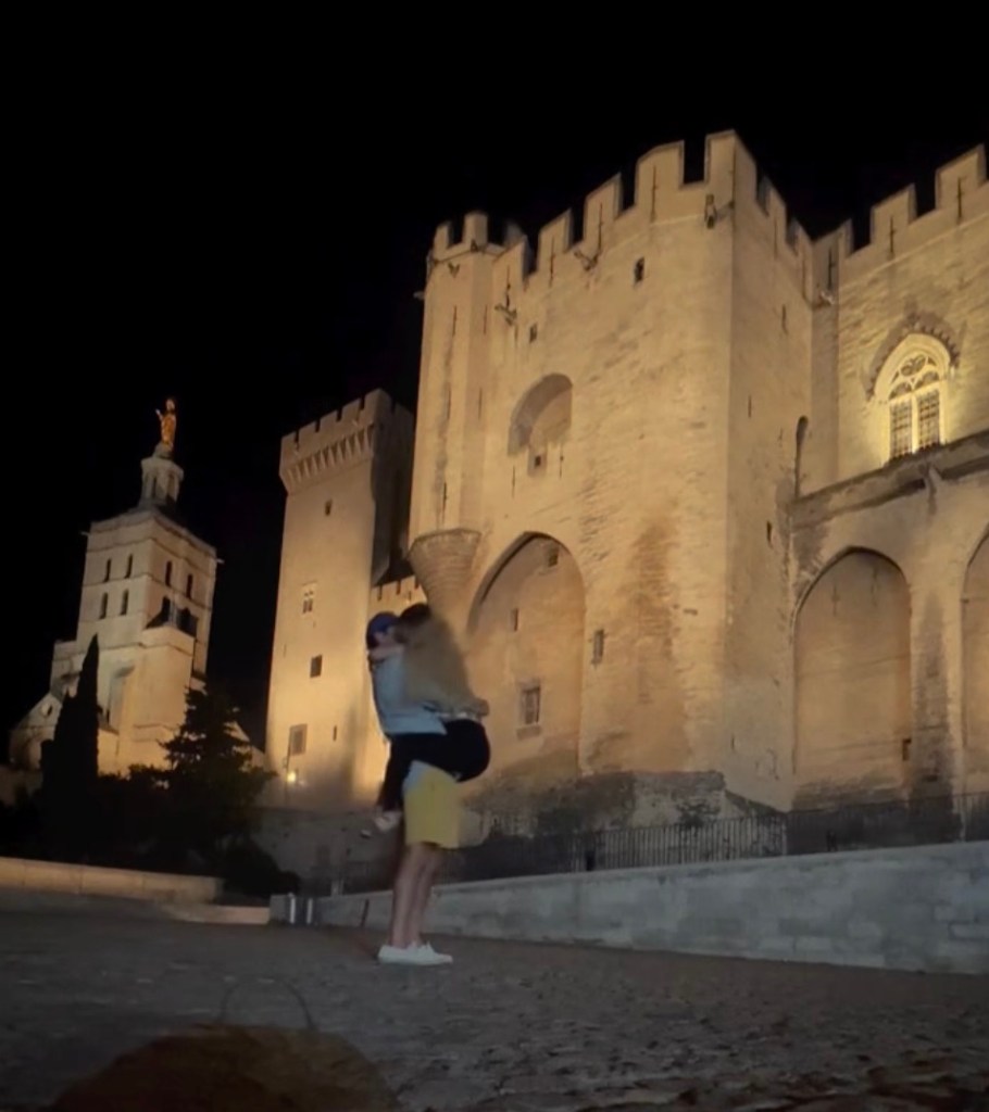 A man holding a woman in his arms, embracing her in front of a castle at night.
