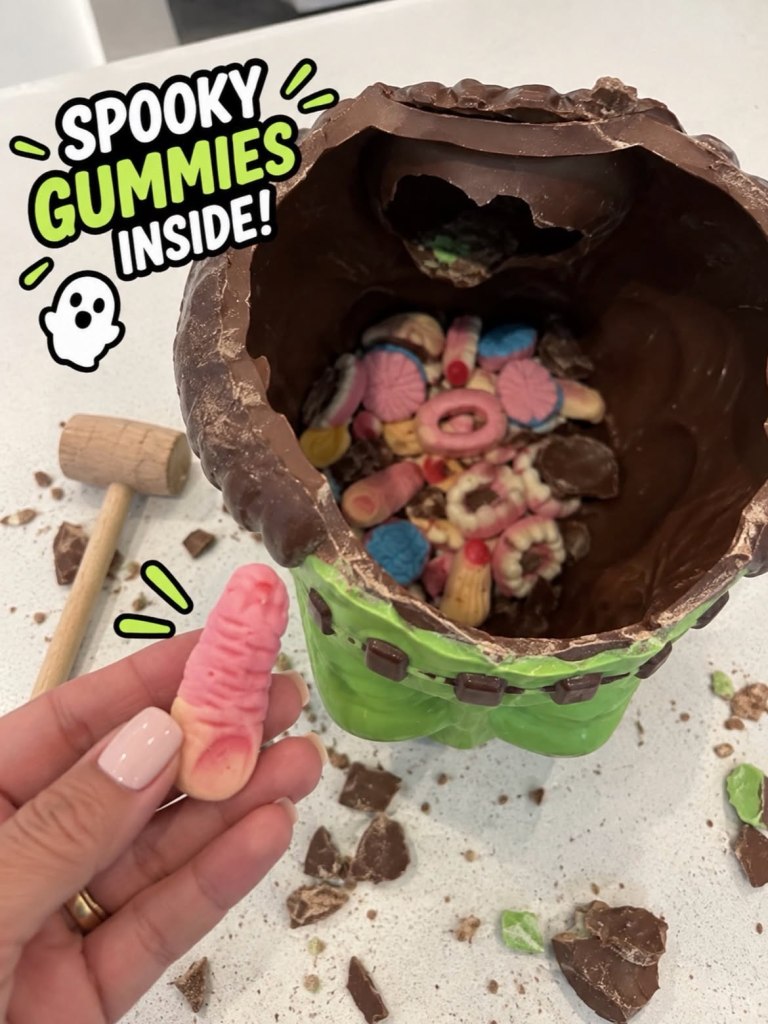 A chocolate Frankenstein head filled with various gummies, with a hand holding a finger-shaped gummy.