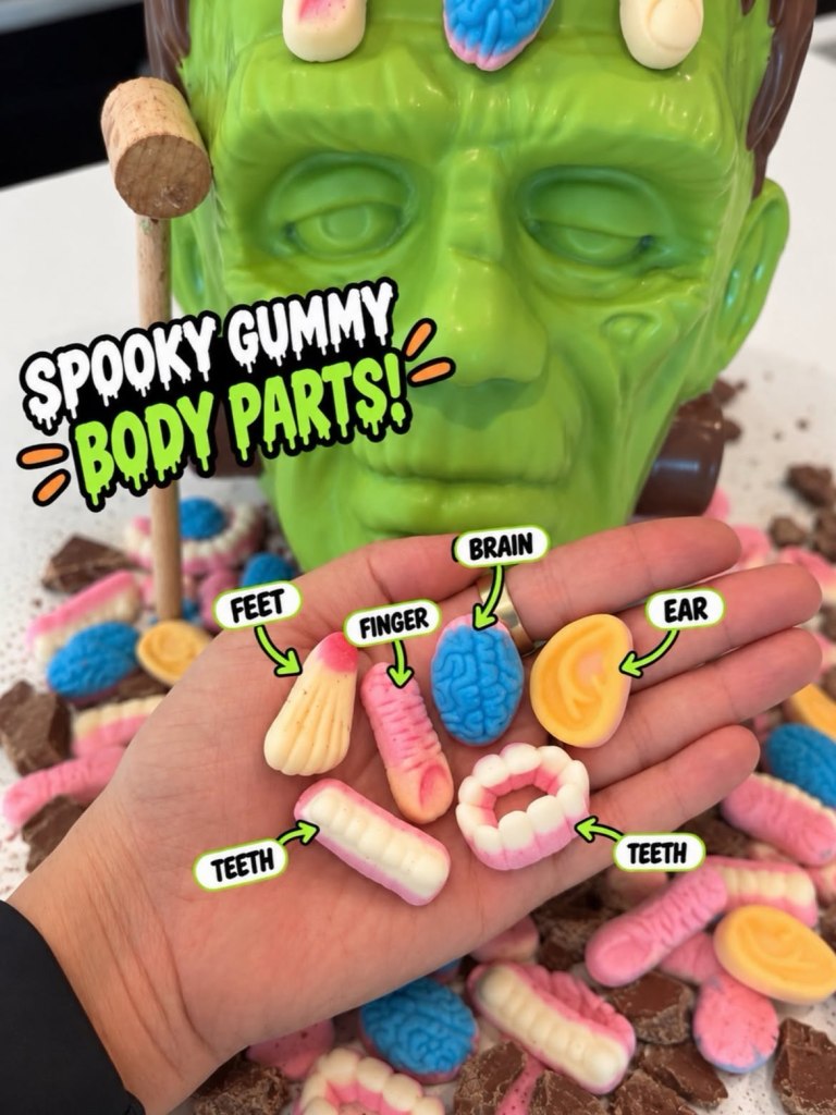 A chocolate Frankenstein head with a hand holding gummy body parts including feet, finger, brain, ear, and teeth.