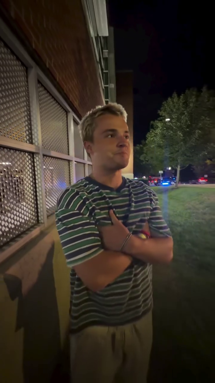 A young man with blonde hair and a striped shirt stands with his arms crossed, looking away from the camera.