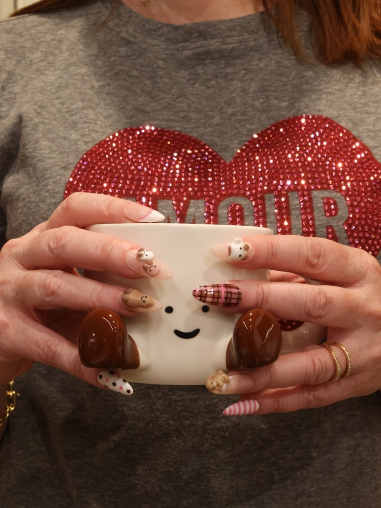 A woman in a gray sweater with a red sparkly heart design holding a white mug with a smiling face and brown feet.