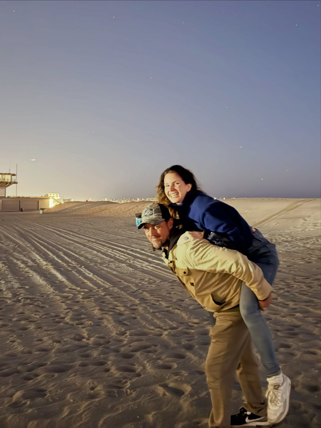 Lana Del Rey getting a piggyback ride from Jeremy Dufrene on a sandy beach at dusk.