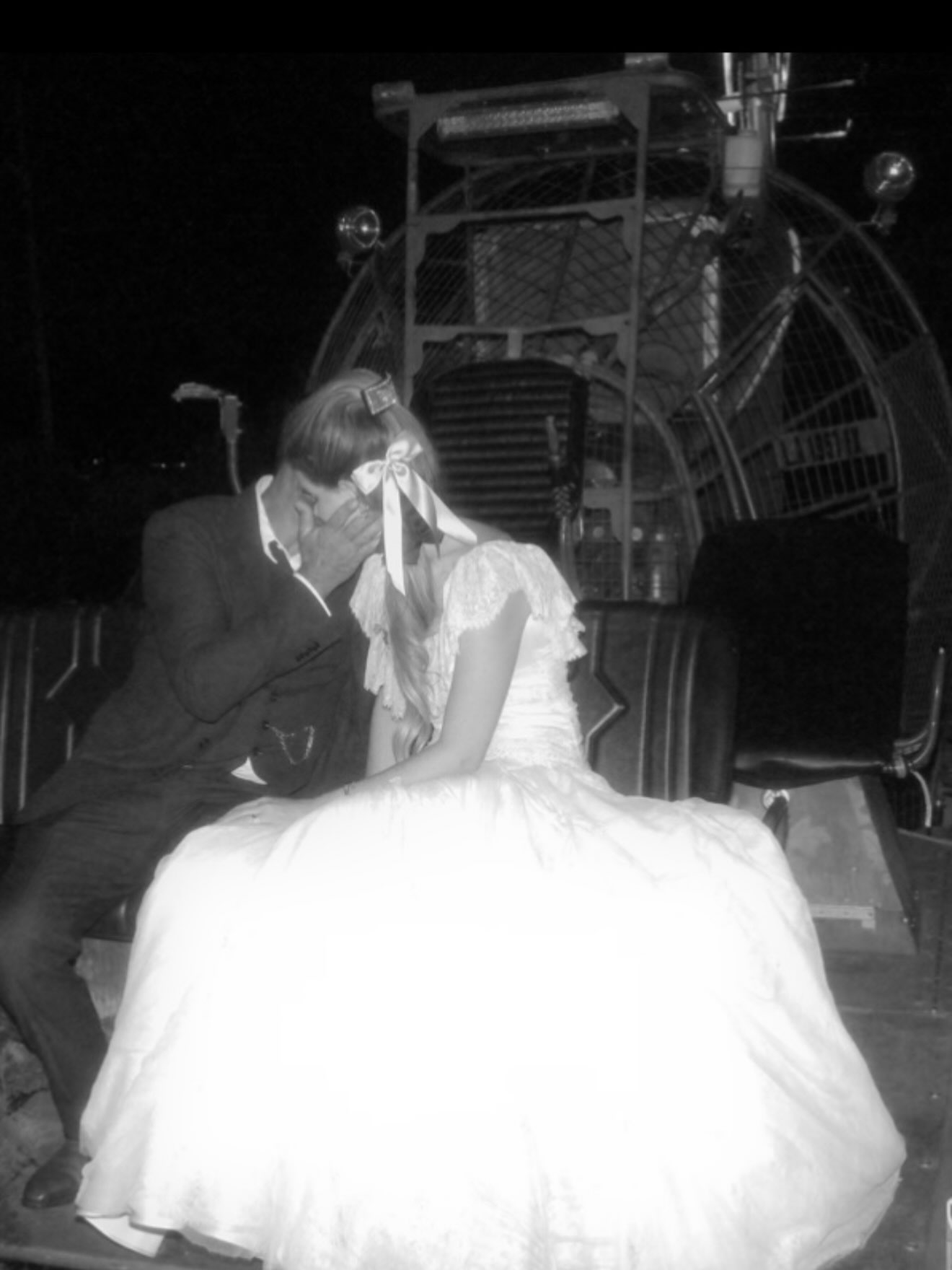 Black and white photo of a man in a suit kissing a woman in a wedding dress in front of an airboat.