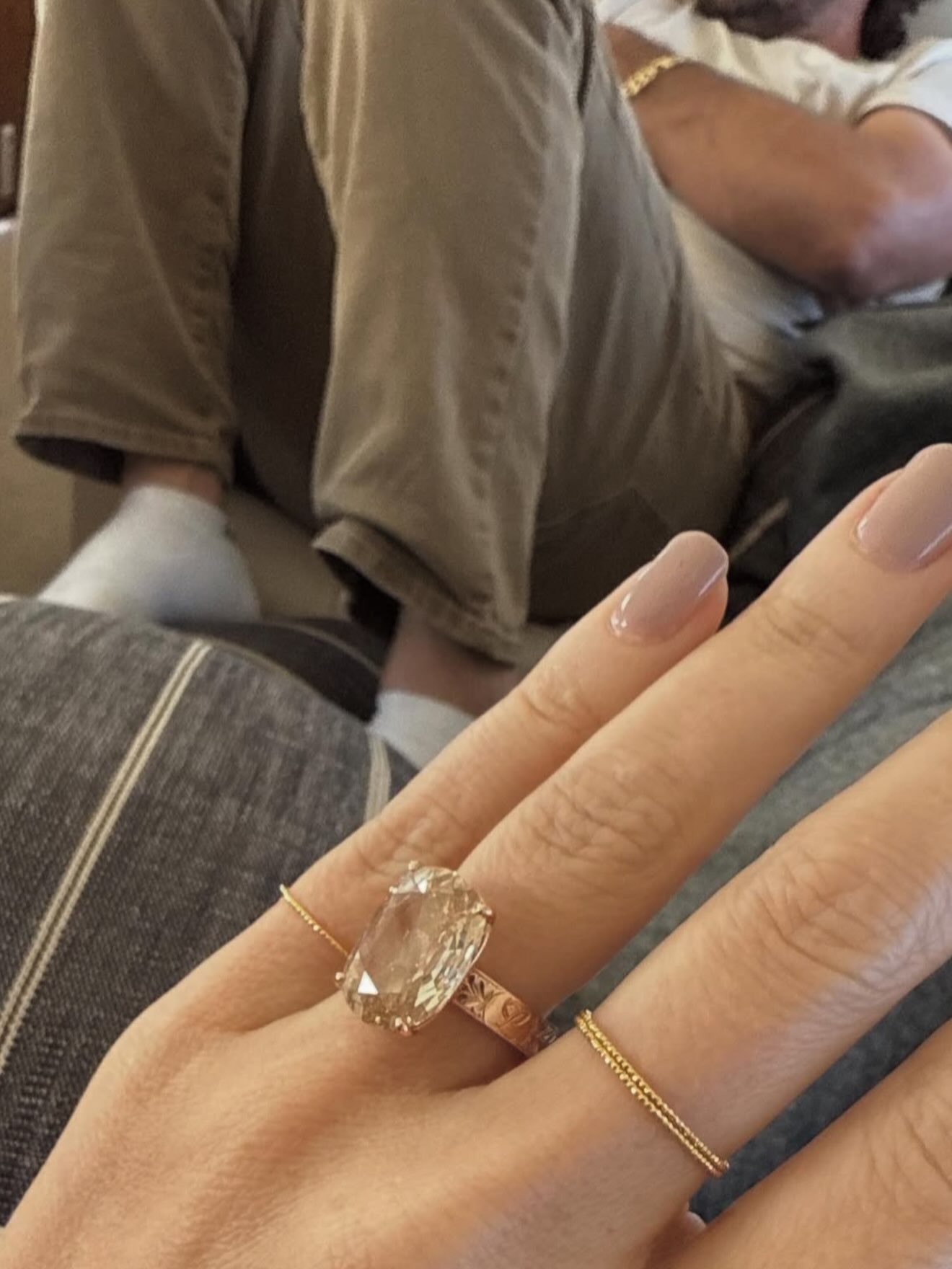 Close-up of a hand wearing a large oval engagement ring and a thin gold band, with a person in khaki pants and a white shirt in the background.