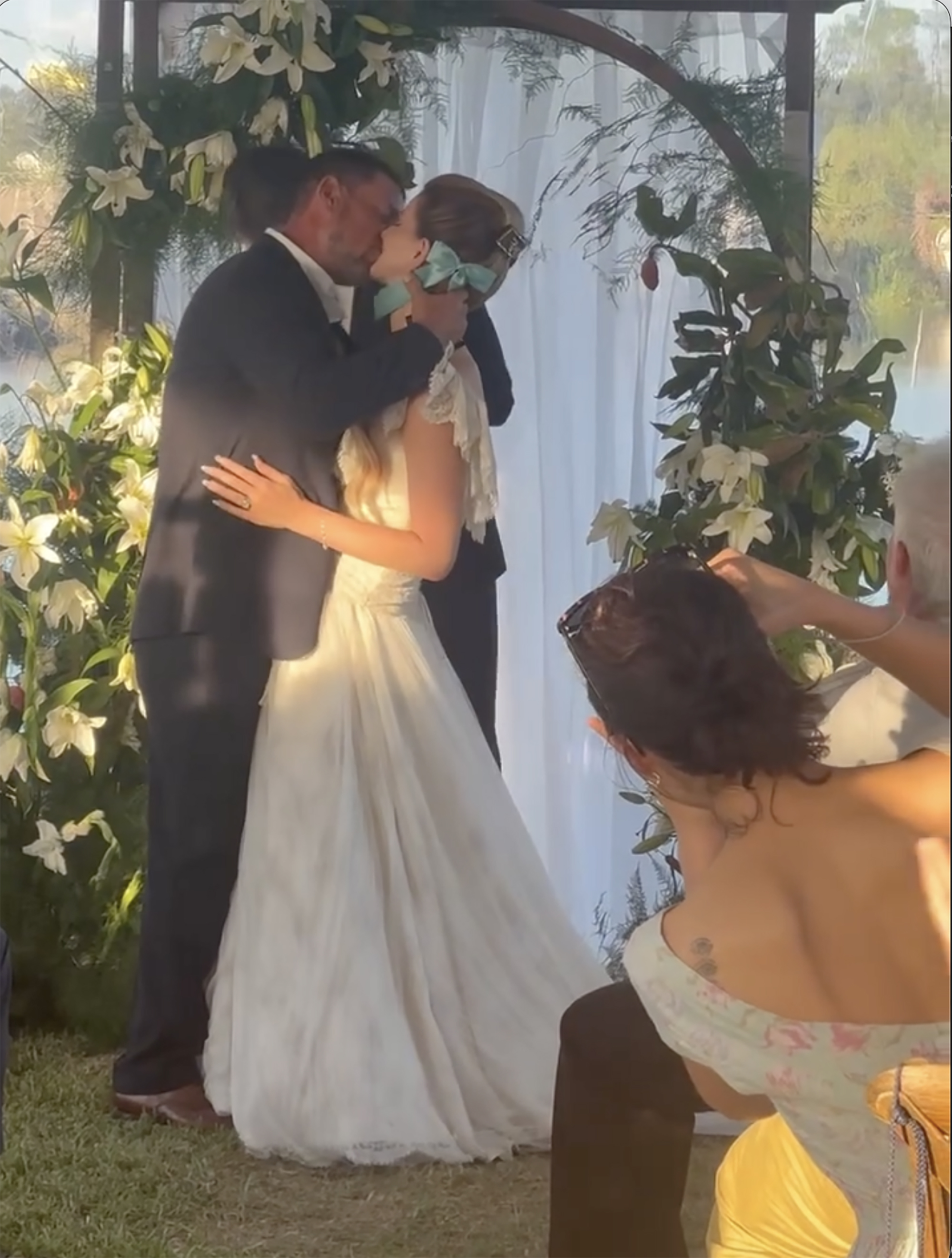 Lana Del Rey, in a white wedding gown, and Jeremy Dufrene, in a black suit, kiss under a floral arch.