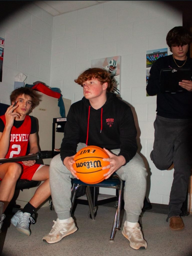 Three young men in a room, two seated and one standing, the middle one holding a basketball.