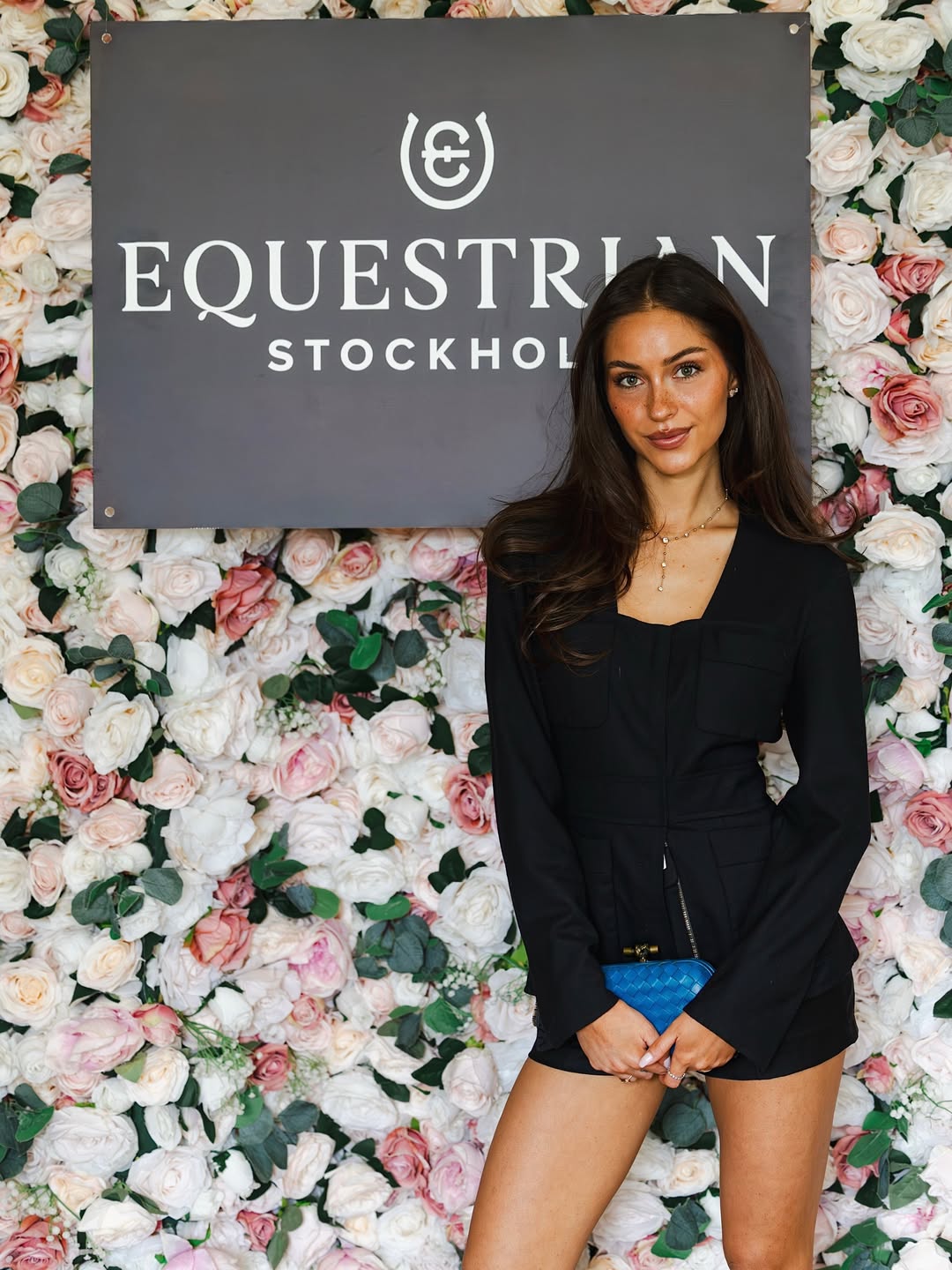 A woman in a black romper stands in front of a flower wall with an 