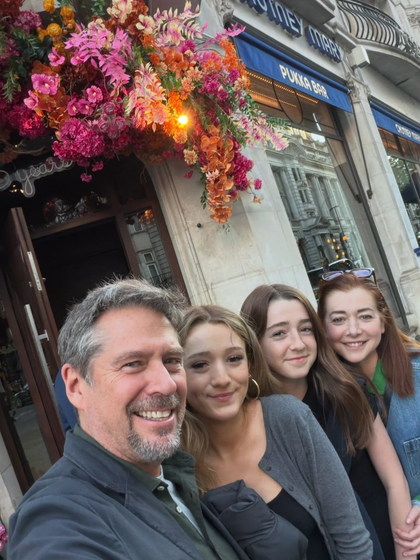 Alyson Hannigan and family posing for a selfie in front of a building with colorful flowers and a sign that says 