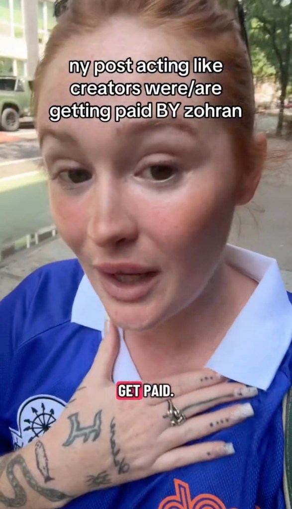 A woman with red hair speaks to the camera, wearing a blue shirt with a white collar. Her right hand, covered in tattoos, rests on her chest. Text overlays read 