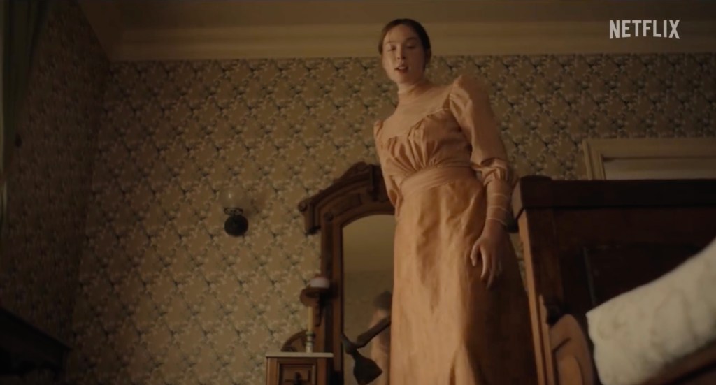 A woman in a peach dress stands in a room with patterned wallpaper, looking down at a bed from the