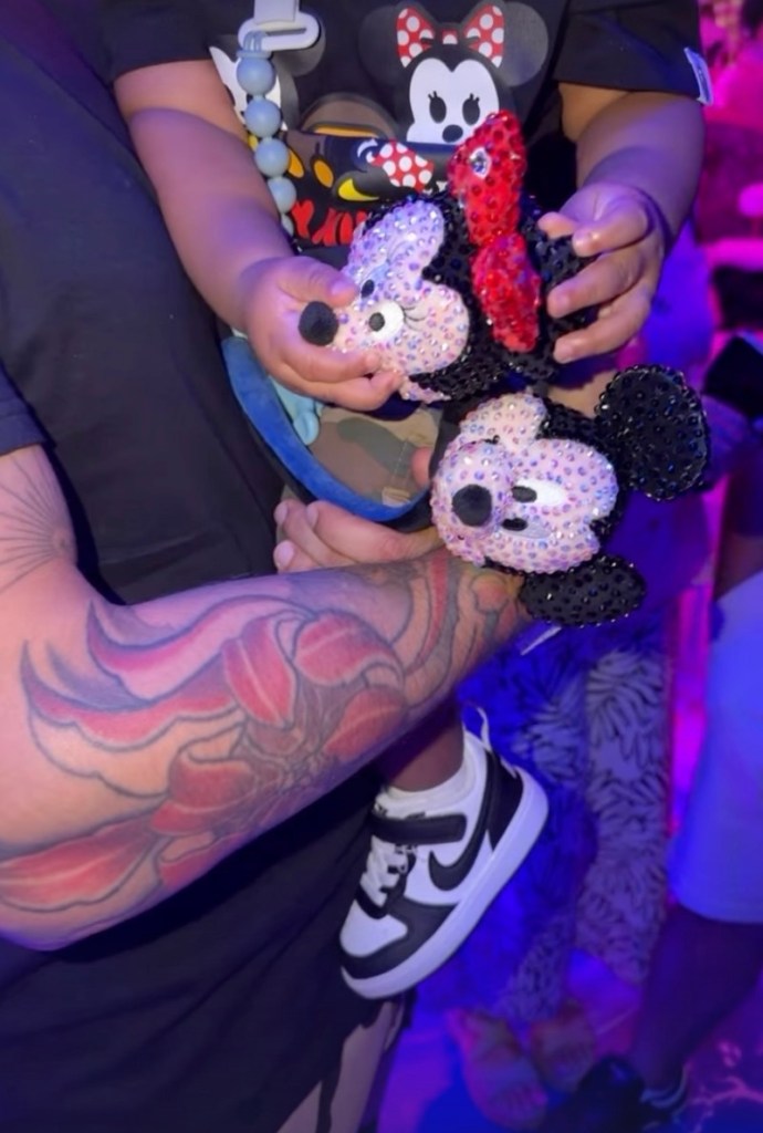 Cardi B and Offset's son, Wave, wearing a Minnie Mouse shirt and holding a Minnie Mouse toy, is being held by Offset, whose tattooed arm is visible.