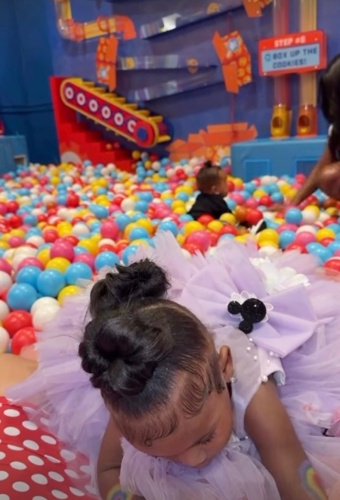 A young girl with her hair in a bun wears a lavender dress in a ball pit.