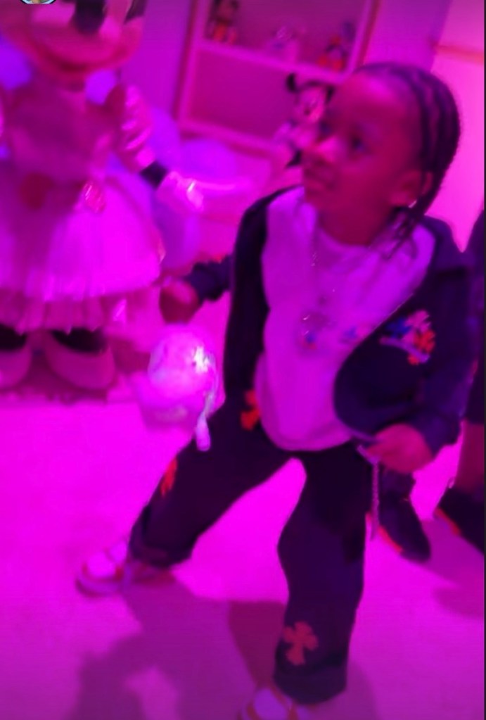 A young child in a dark tracksuit with colorful cross motifs, holding a glowing toy in a room lit with magenta light.