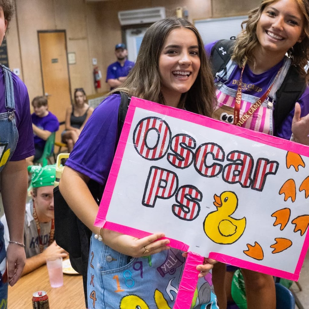 Two smiling young women in purple shirts and overalls hold a handmade sign that reads 