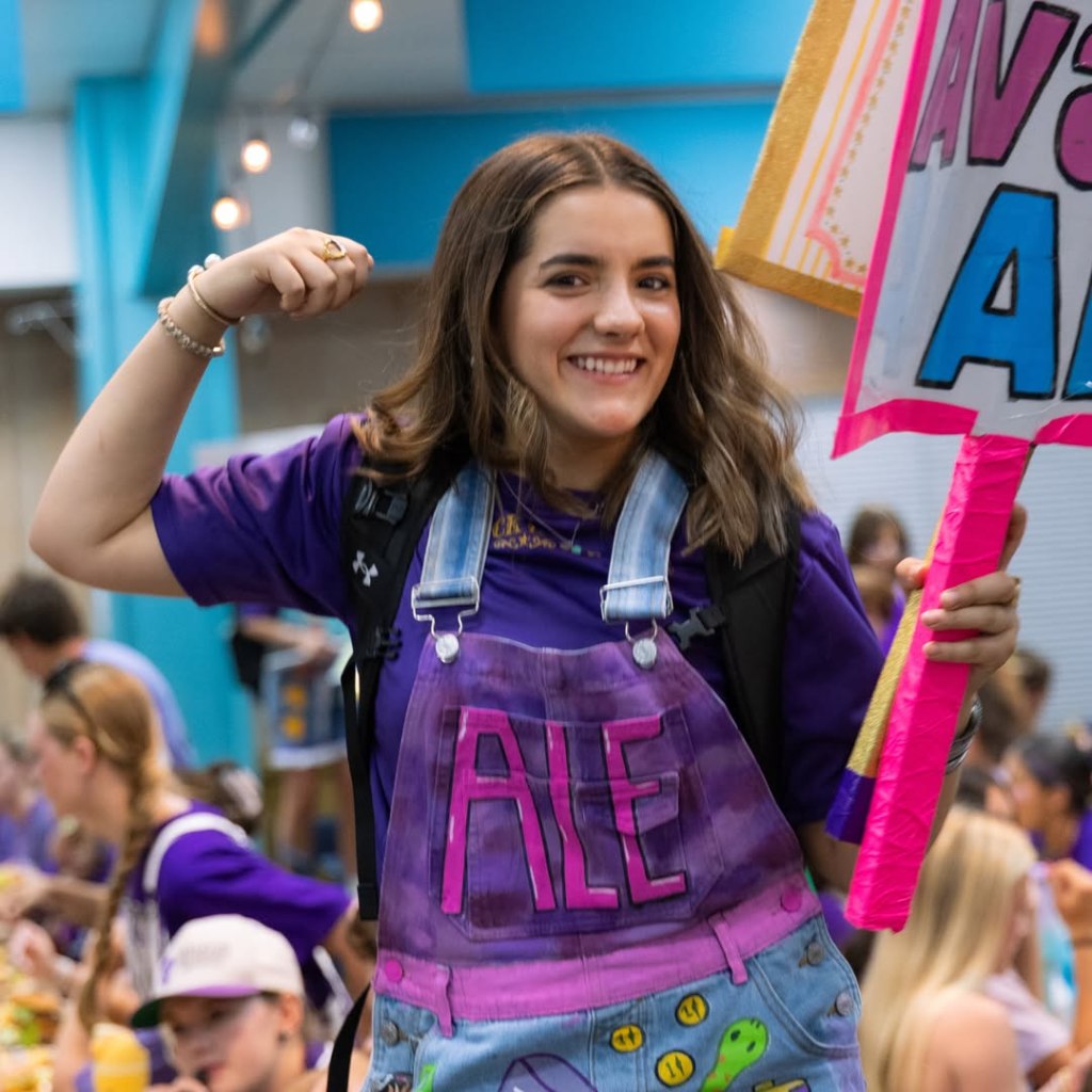 A young woman smiling, holding a sign, and wearing a purple shirt under personalized denim overalls that say 