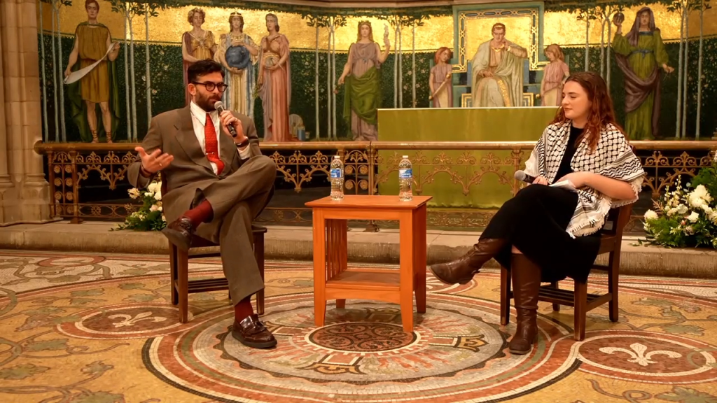 A man holding a microphone and a woman wearing a keffiyeh sit on chairs facing each other on a patterned floor, with a golden altar behind them.