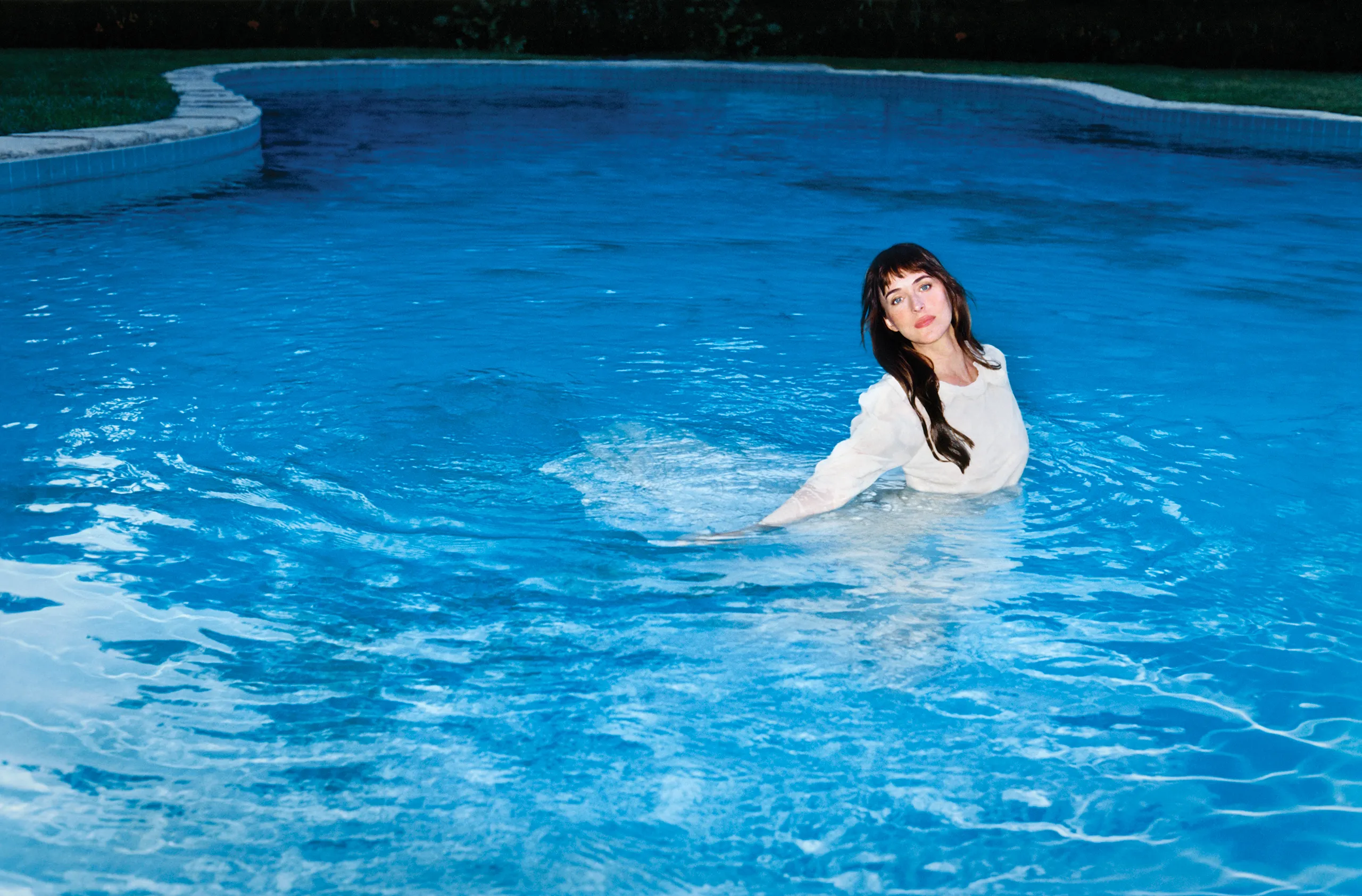 Dakota Johnson in a white dress in a swimming pool, looking at the viewer.