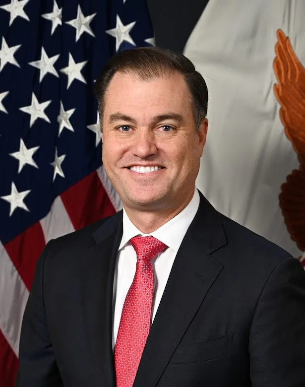 Headshot of Macon Dane Hughes smiling, wearing a dark suit and red tie, with an American flag and a flag with an eagle visible in the background.