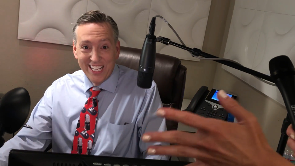 Jim Thornton in a blue shirt and red tie smiling into a microphone.