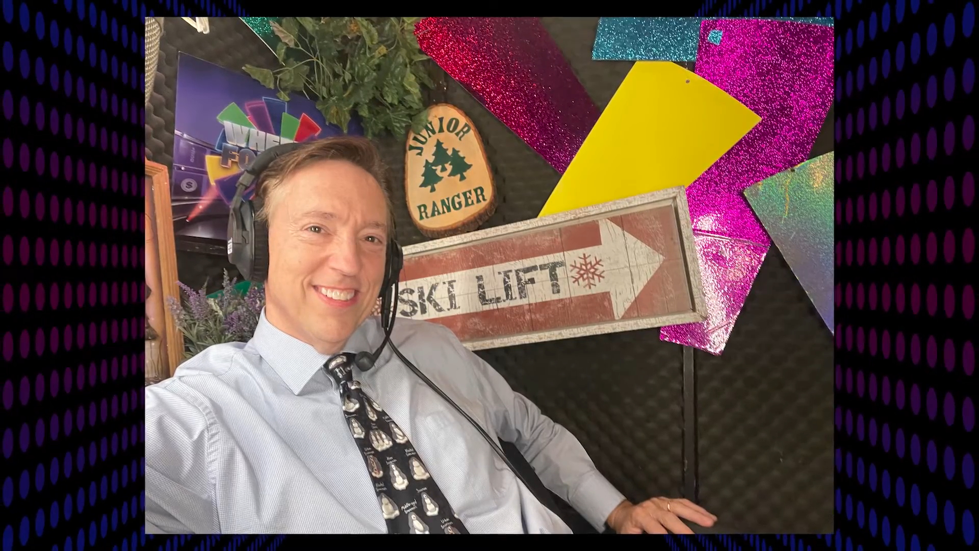 Jim Thornton, a Wheel of Fortune announcer, smiles for a selfie while wearing a headset and a tie with penguin characters, sitting in front of colorful decor.