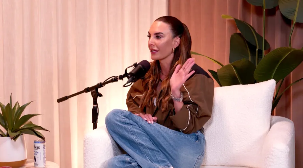 Elizabeth Chambers speaking into a microphone with her hand raised, wearing a brown shirt and jeans.