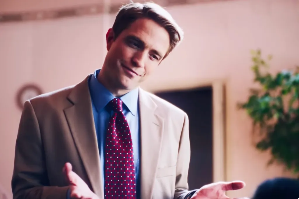 A man in a tan suit, blue shirt, and red patterned tie shrugs with his hands open.