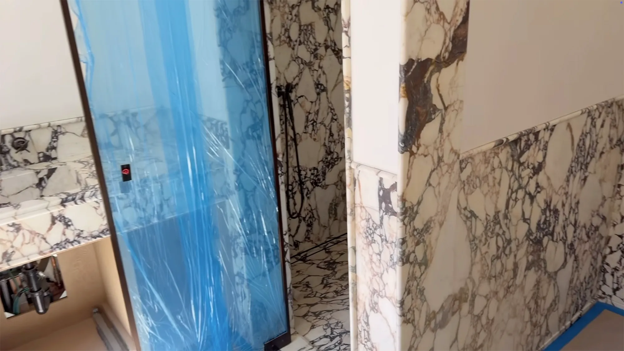 A bathroom with marble walls and a shower covered in blue protective film, under construction.