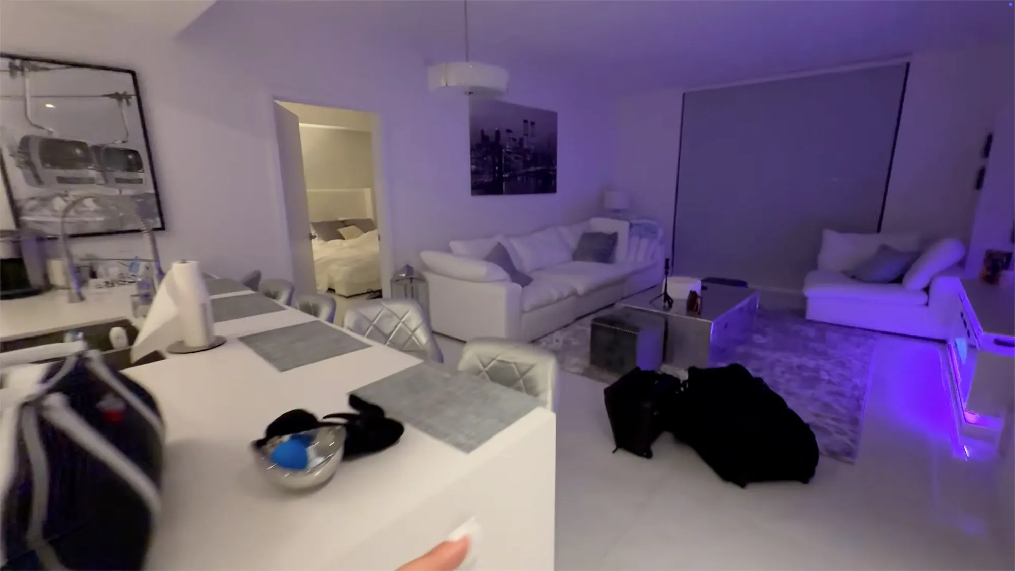 A wide-angle view of a modern college apartment interior with white walls, white furniture, a kitchen island, and purple lighting.