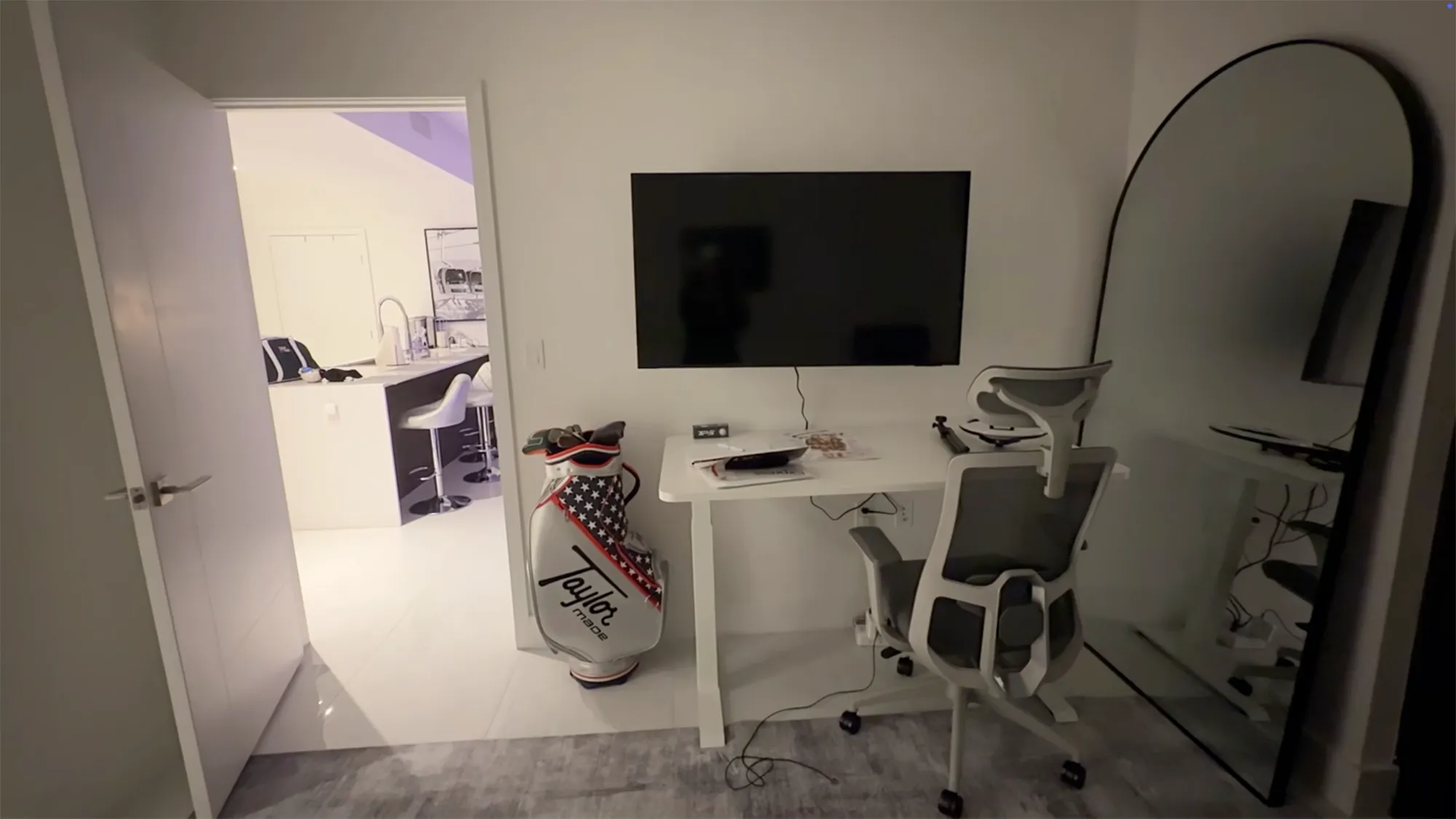 A white college apartment with a white kitchen, a study area with a TV, a white desk and a chair, and a white golf bag.