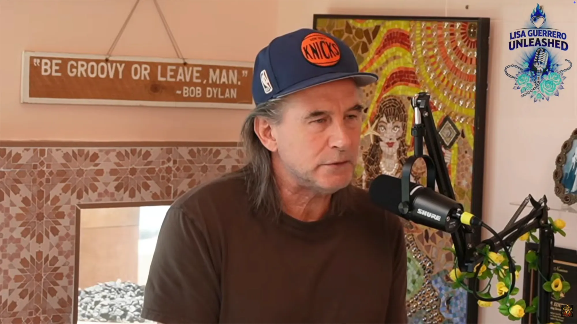 Billy Baldwin with long hair and a Knicks baseball cap speaks into a Shure microphone.