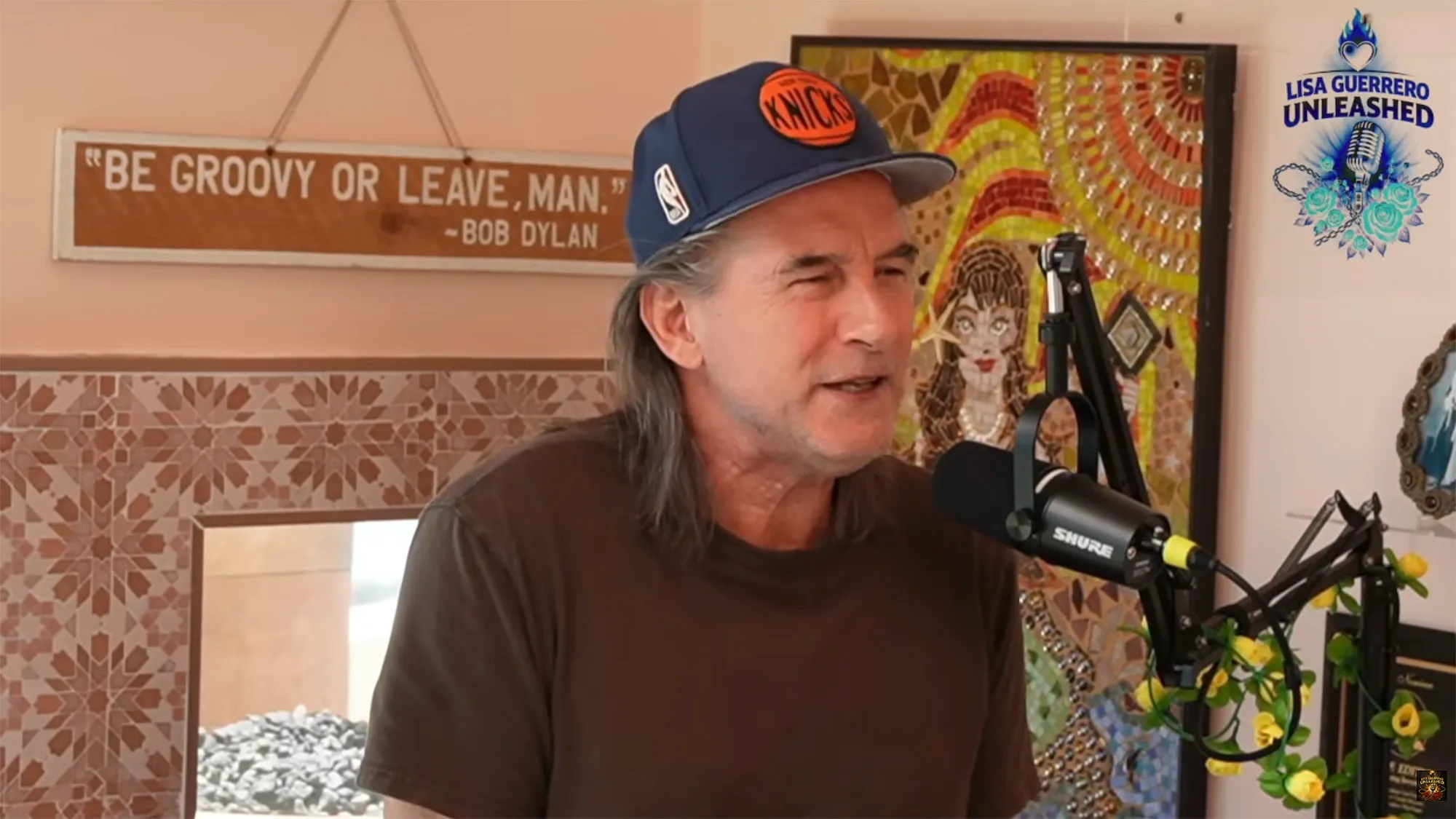 Billy Baldwin wearing a Knicks cap and a brown shirt while speaking into a Shure microphone.