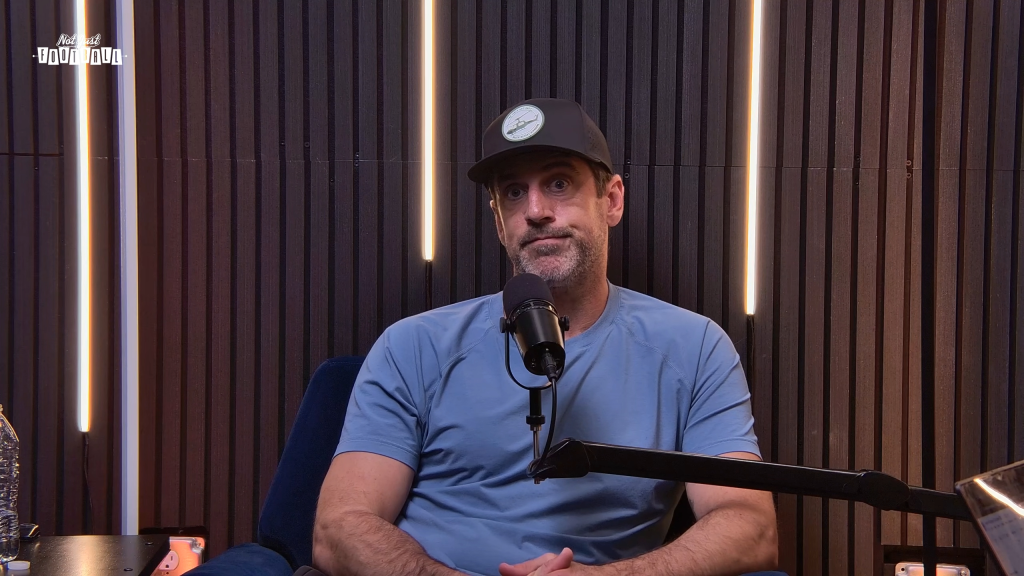 Aaron Rodgers wearing a baseball cap and light blue shirt, speaking into a microphone during an interview.
