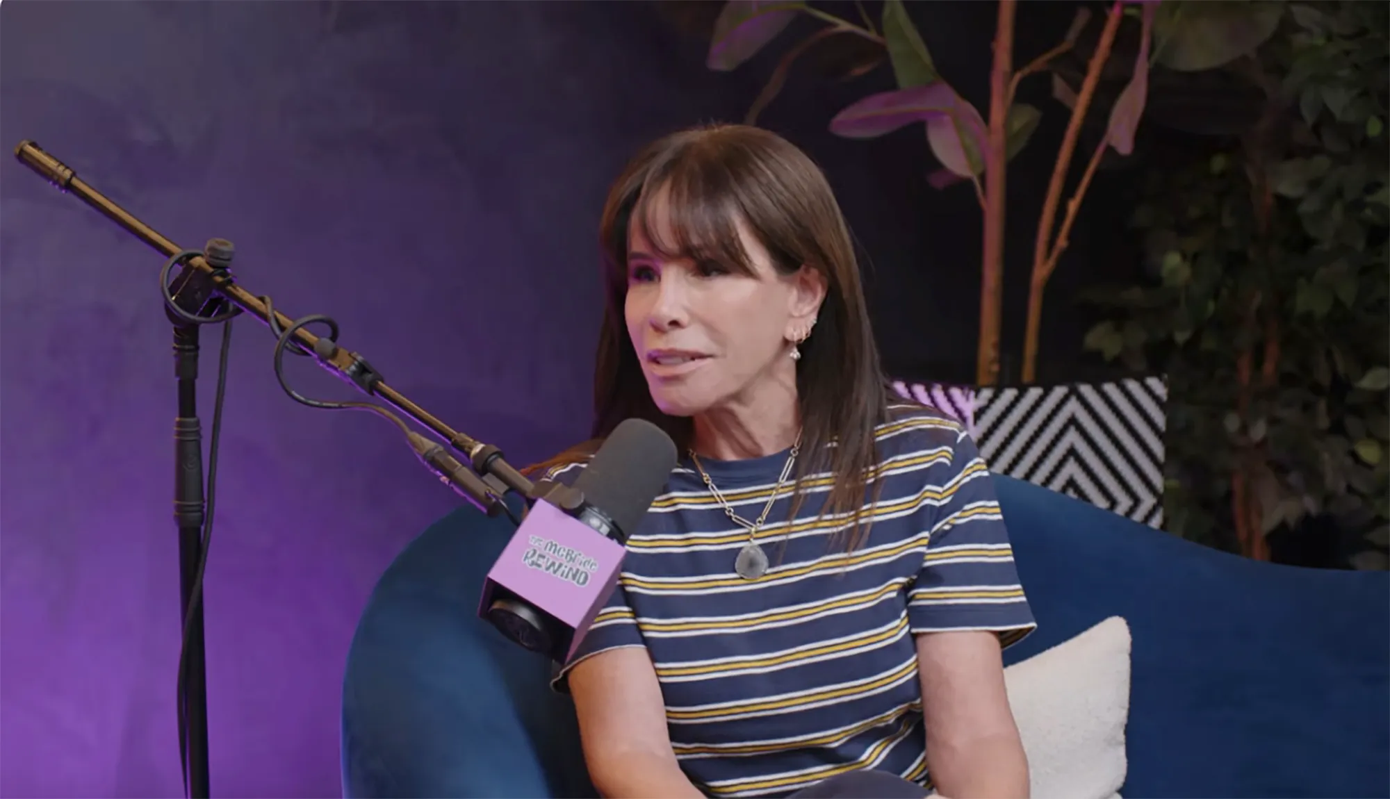 A woman with long brown hair and bangs, wearing a striped shirt and a necklace, speaks into a microphone labeled 