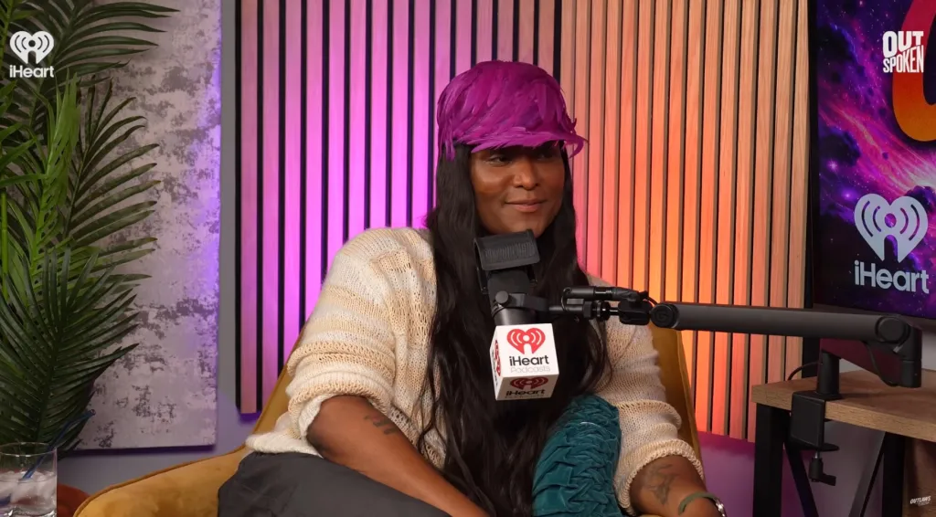 Law Roach in a purple feathered hat sitting in front of a microphone.