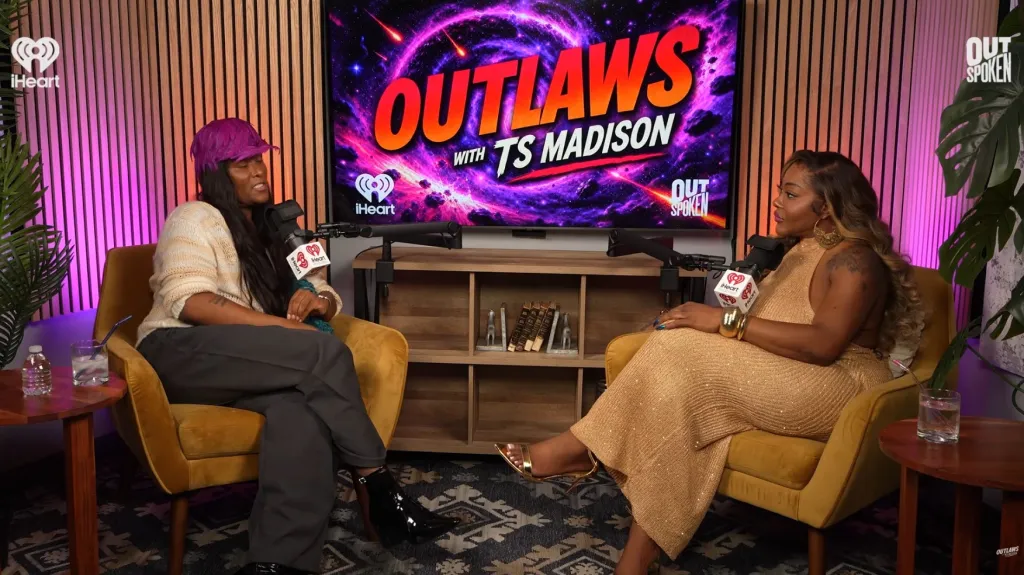 Two women, TS Madison on the right in a gold outfit and another woman on the left in a purple hat, sit opposite each other during a podcast recording for