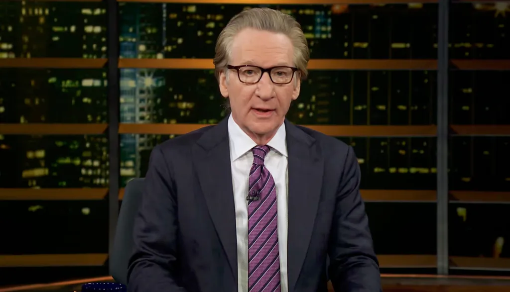 Bill Maher sitting in a suit and tie against a blurred city background at night.